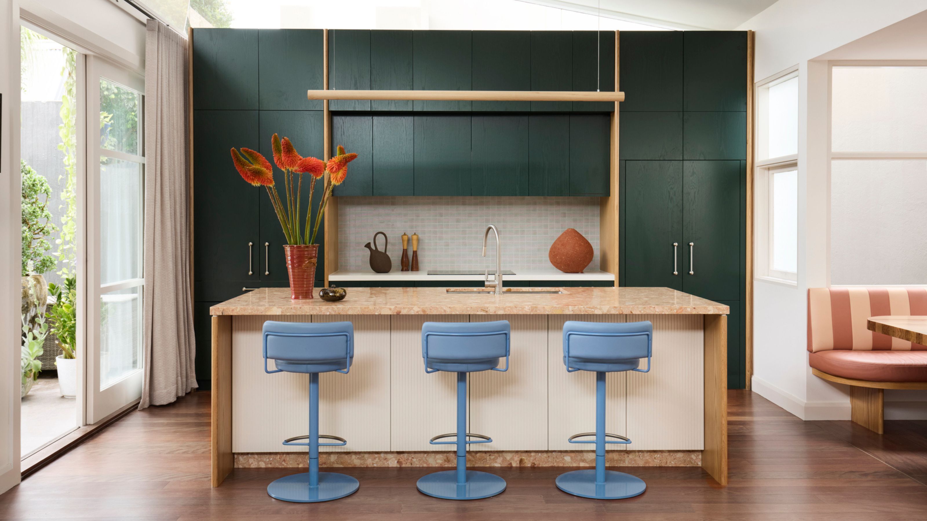 A colorful kitchen with dark green cabinets and blue bar stools in front of an orange marble island with a vase of fluffy vibrant flowers on top next to open sliding doors