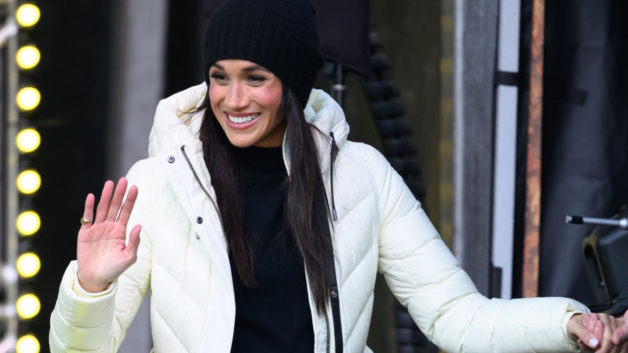 Meghan Markle wearing a white puffer coat and black hat waving and smiling on stage 