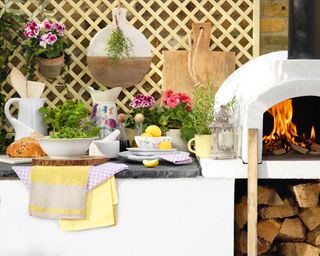 A white outdoor kitchen with yellow textiles and crockery, potted herbs and pink flowers, and a wooden trellis in the background.