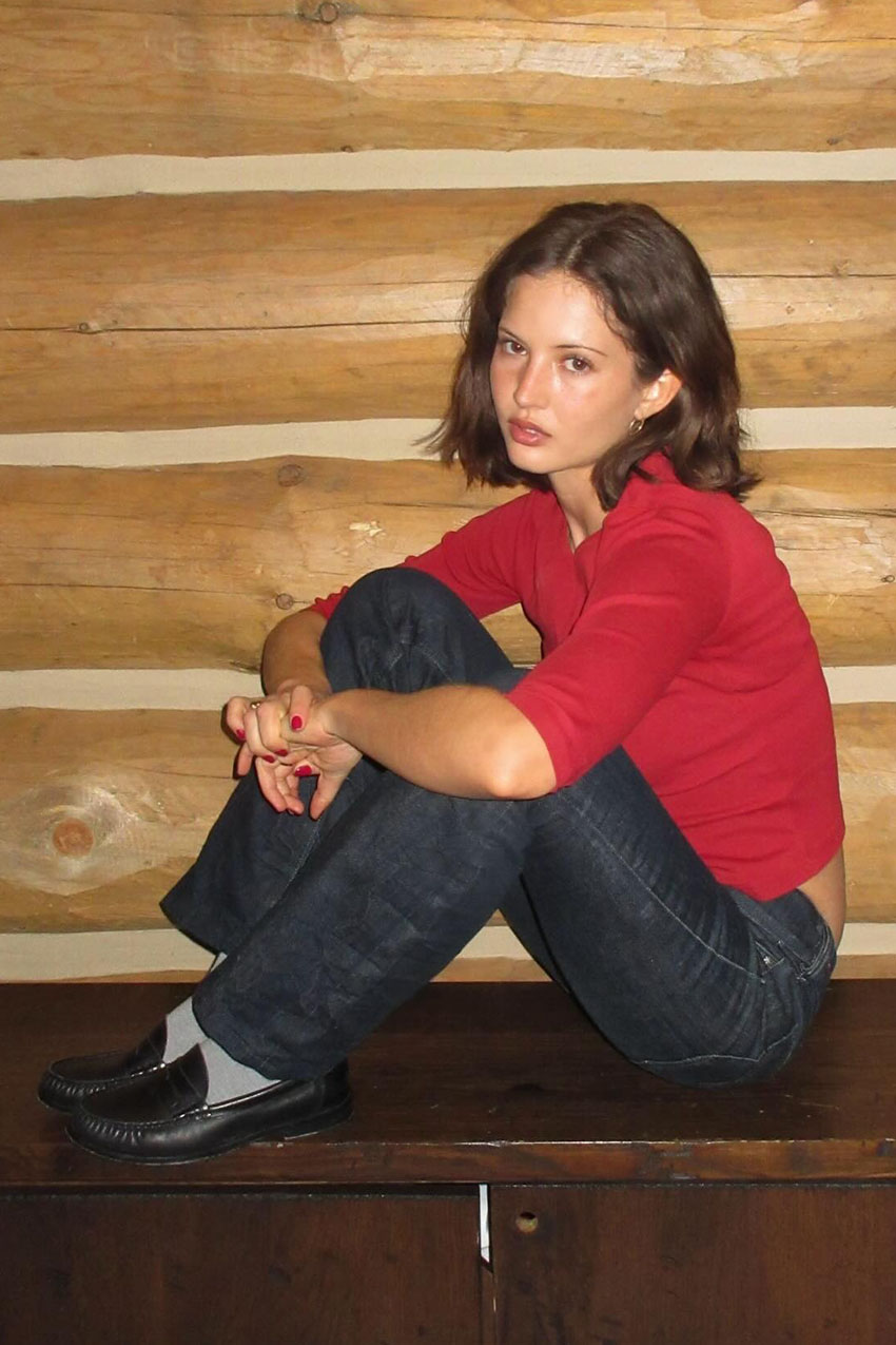 A woman wearing a red long sleeve top, dark blue jeans, grey socks, and black loafers