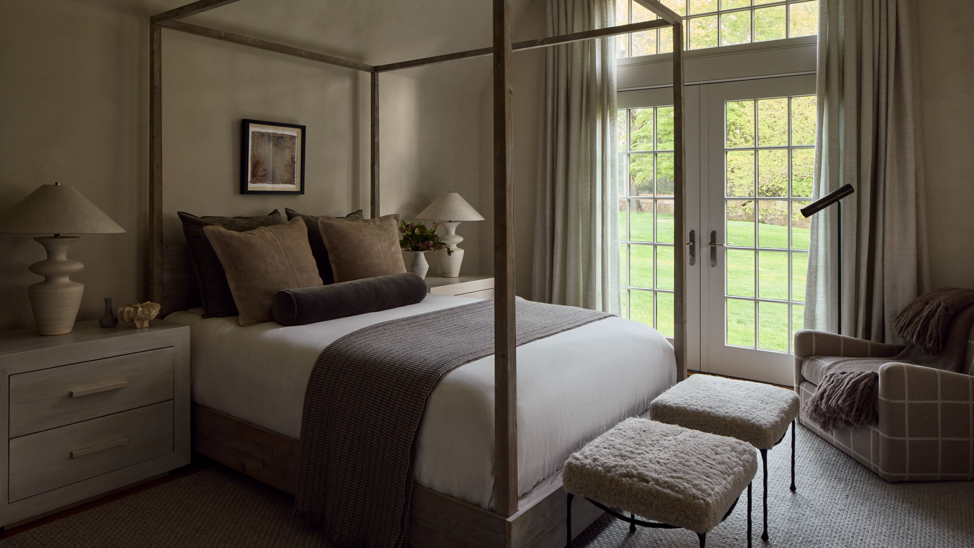 Neutral bedroom with four poster bed and large French windows. There are long drapes around the windows and the bed is styled with layered of neutral sheets