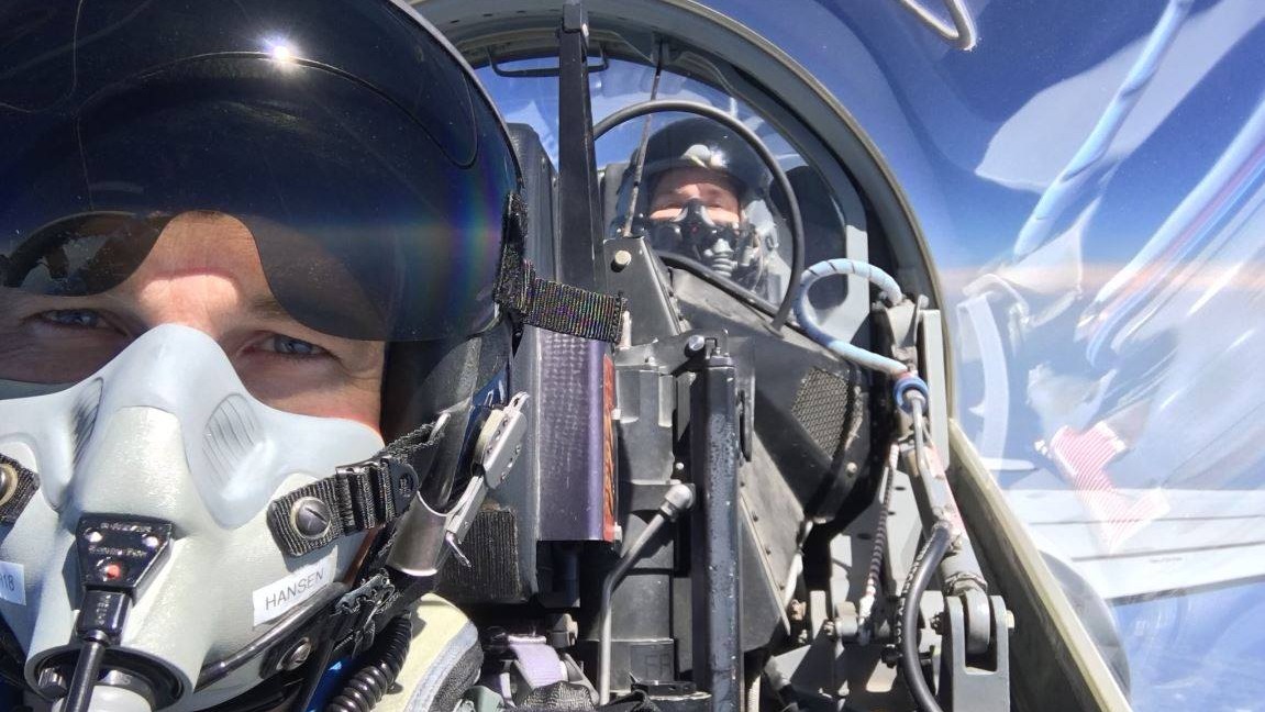 a cockpit view of jeremy hansen masked up with sunglasses and an oxygen mask. behind him is the dome of the aircraft and another astronaut, jenni sidey-gibbons, barely visible in another seat