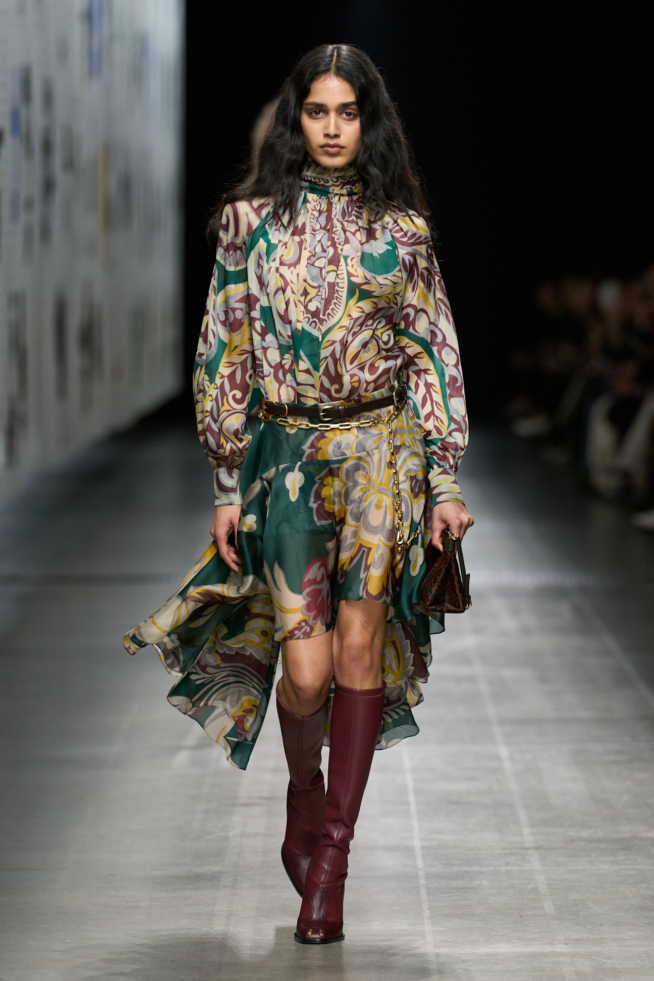 Model walking in the Etro 2025 runway show wearing a jewel-toned mini boho dress, slim black belt and burgundy knee boots.