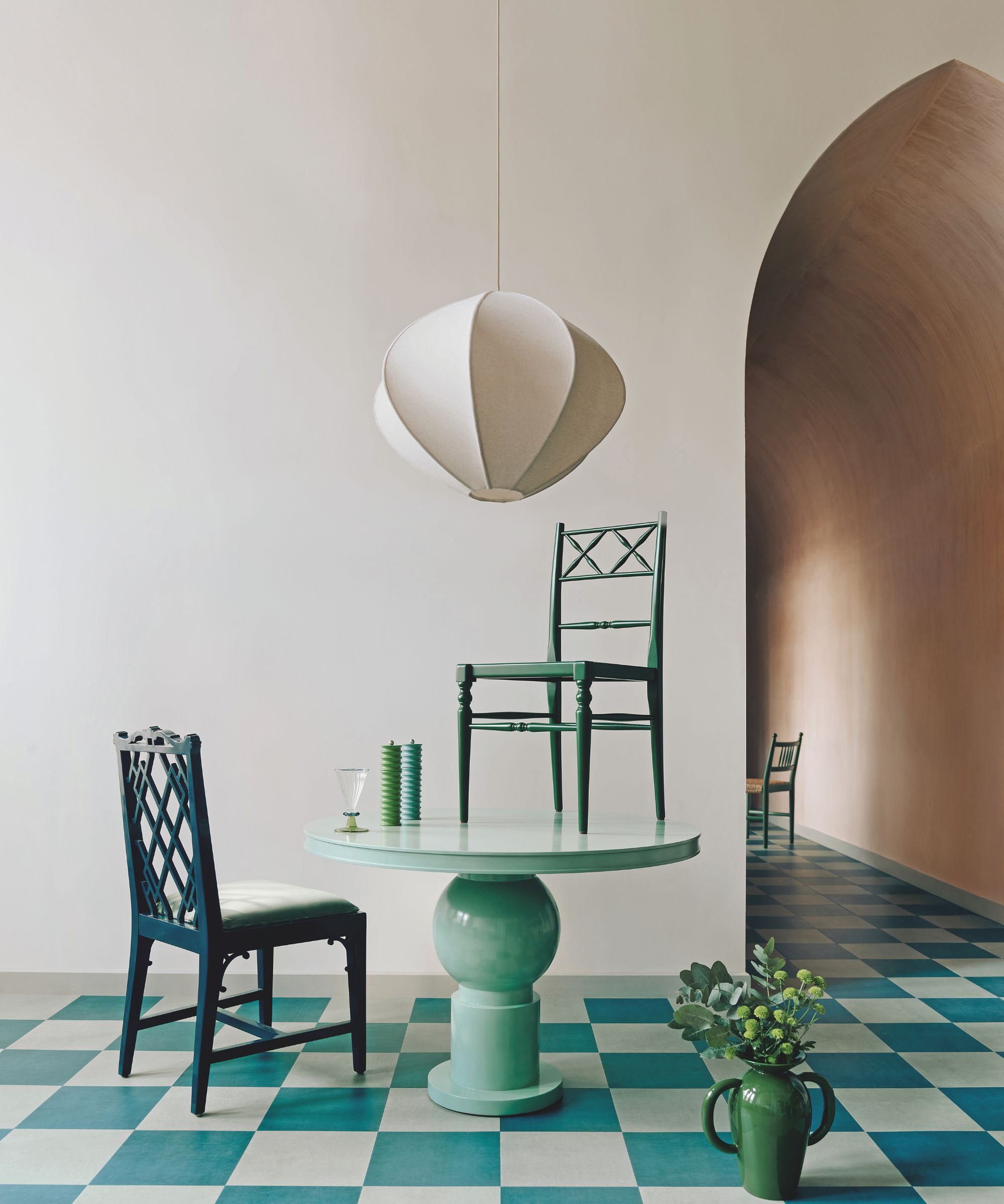 A surrealist dining area with a mint-green pedestal table and a matching green chair perched on top. The room features teal-and-white checkered flooring and a sculptural, white pendant lamp hanging above.
