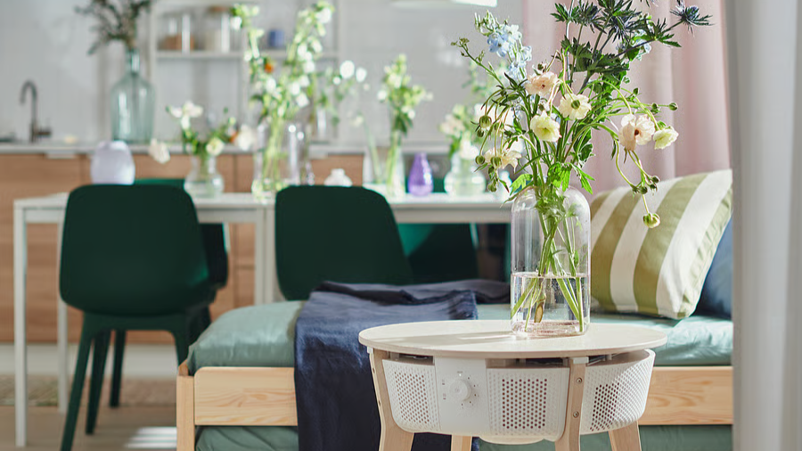 A brightly lit room with flower vases on the table, green chairs and a bed with colorful bedding