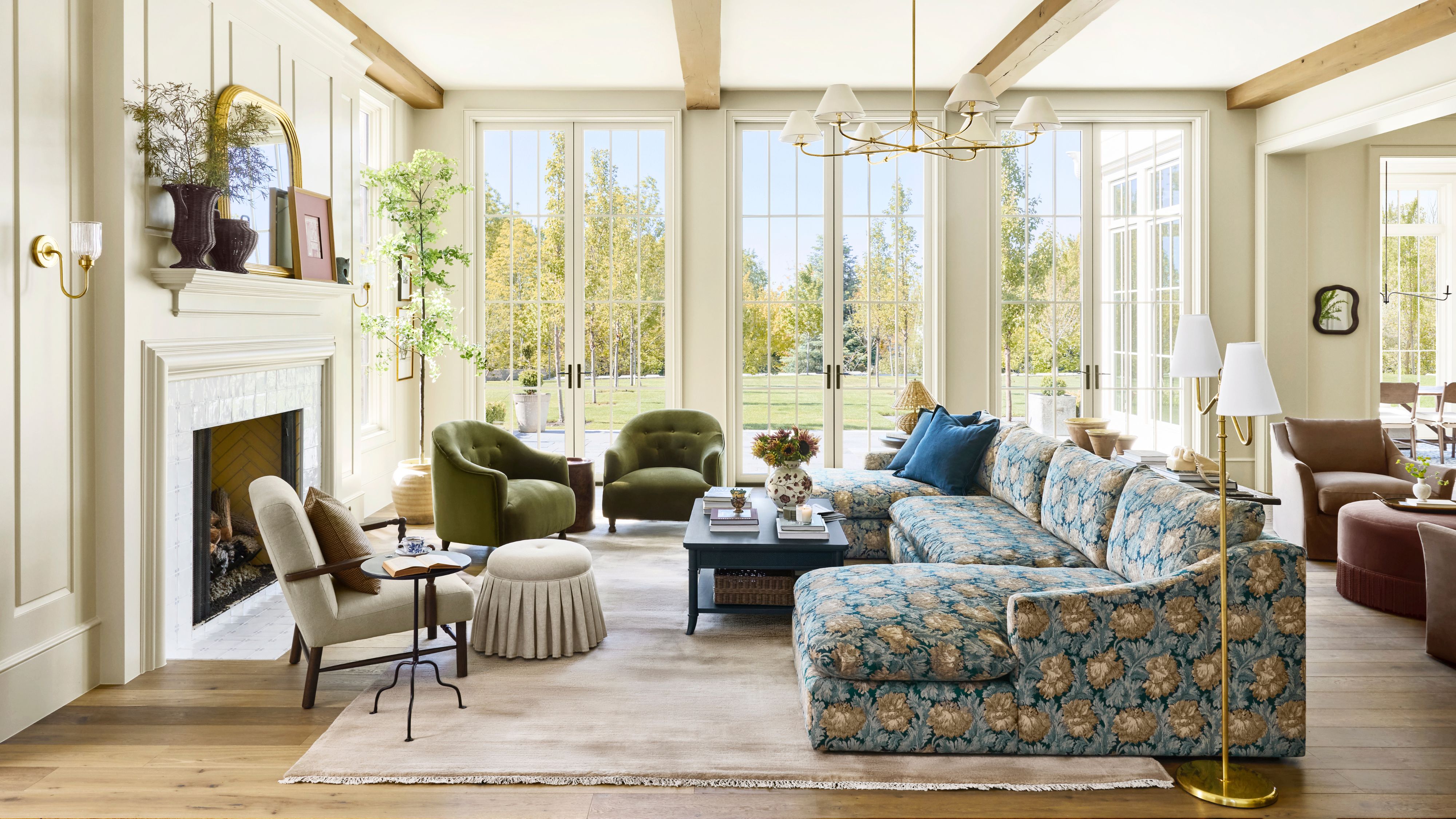 Cheerful sun-filled living room with large windows styled with skirted accent furniture, a neutral rug, a two-shade floral lamp, and a large blue floral sectional.