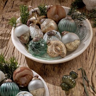 A white bowl place on top of a wooden table, filled with a mixture of metallic and glass baubles