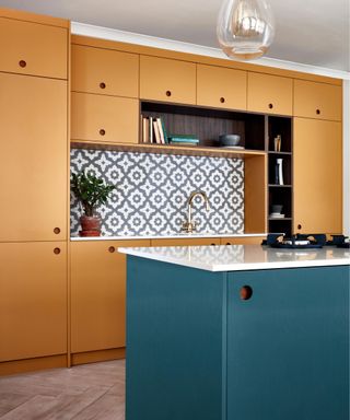 A teal blue kitchen island with a white countertop on top, with mustard yellow cabinets around the wall behind it and a gold and glass pendant light hanging above it