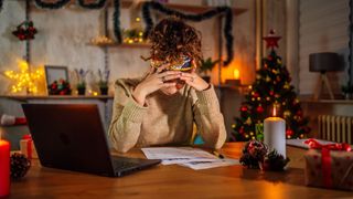 The image shows a dark-haired woman sat to a kitchen table with her laptop open, a notebook on the table in front of her and her head in her hands. Behind her, festive decorations twinkle.