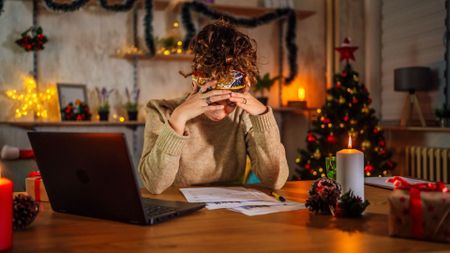 The image shows a dark-haired woman sat to a kitchen table with her laptop open, a notebook on the table in front of her and her head in her hands. Behind her, festive decorations twinkle.