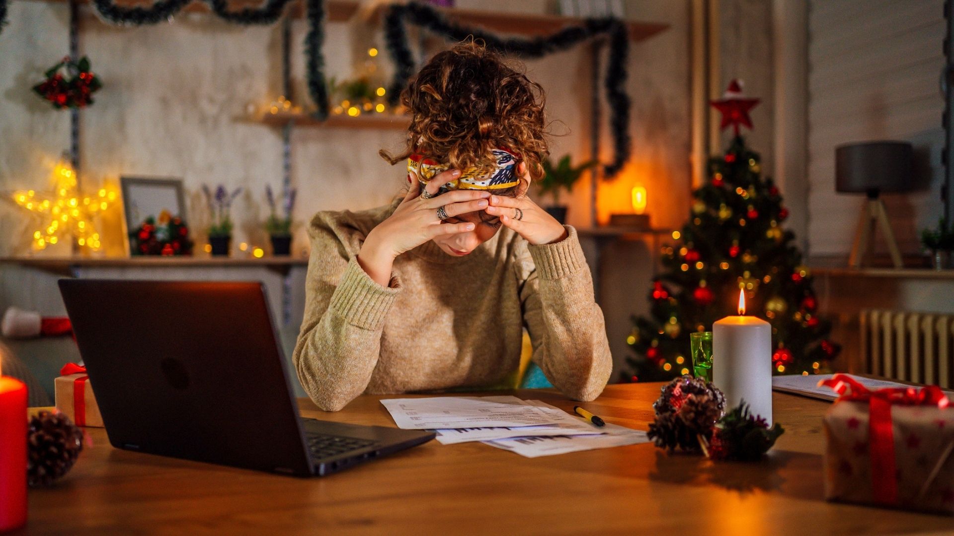 The image shows a dark-haired woman sat to a kitchen table with her laptop open, a notebook on the table in front of her and her head in her hands. Behind her, festive decorations twinkle.