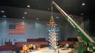 a crane positions a satellite on a test stand inside a large building with an american flag on the wall