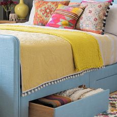 A blue fabric bed with drawers, a mustard coloured throw and bright cushions