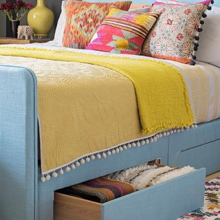 A blue fabric bed with drawers, a mustard coloured throw and bright cushions