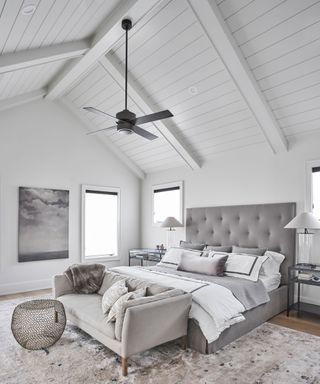 Bright, white bedroom with wooden paneled vaulted ceiling, large bed with gray upholstered frame, white bedding, gray sofa at foot of the bed, black fan light above bed, wooden flooring, gray rug
