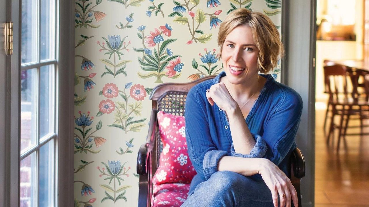 Designer Erin Napier wearing denim sitting in a vintage chair in a room with floral wallpaper