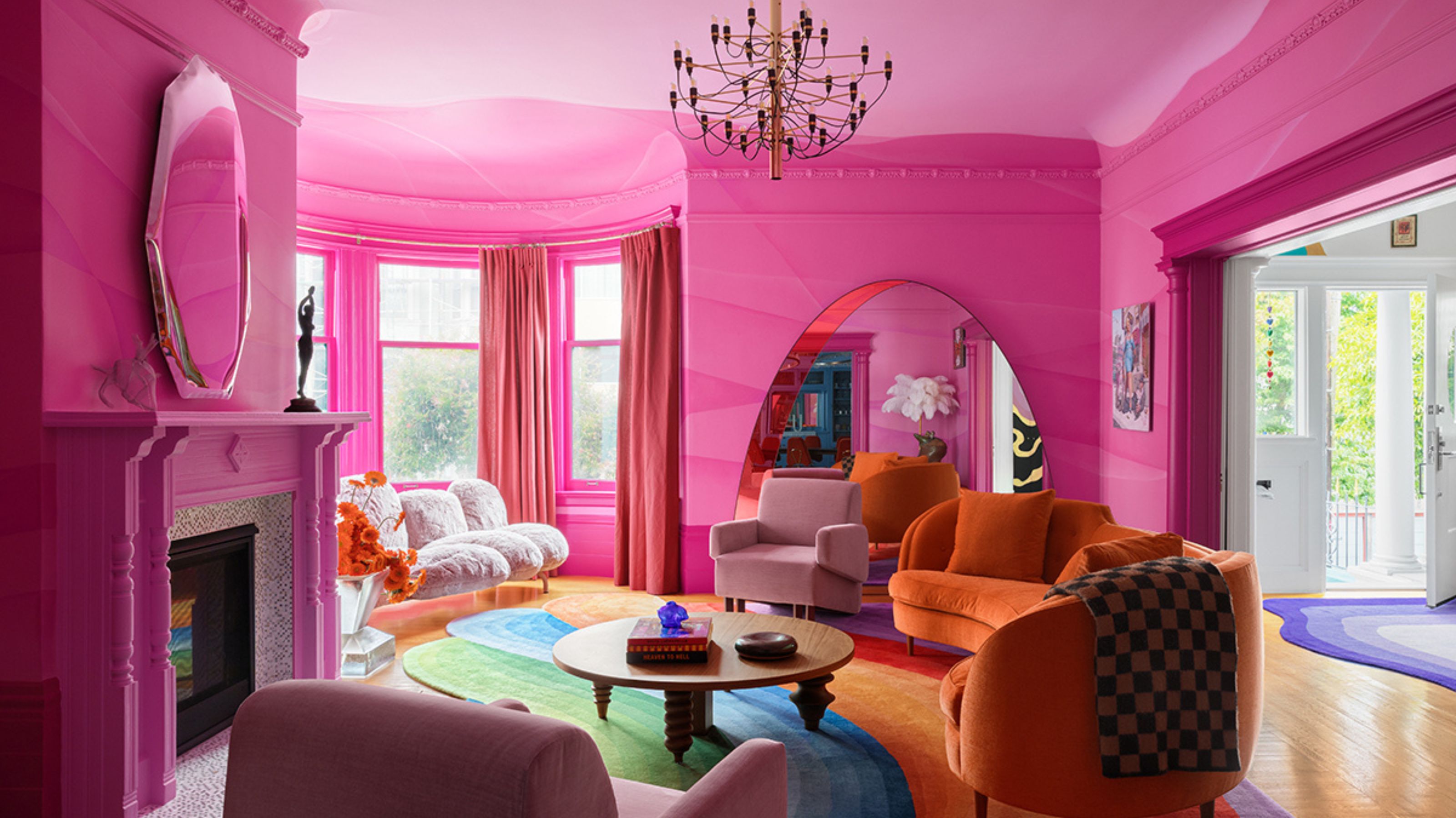 colorful living room with pink color-drenched gradient walls, orange curved sofa, rainbow rug, window seat with fluffy sofa, round timber coffee table, fireplace with mirror above, and timber floors