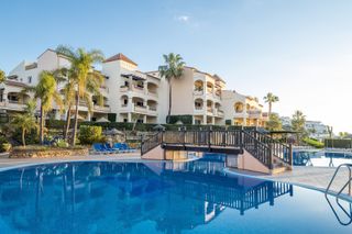 Wyndham Residences, Costa Del Sol. An external shot of one of the apartment buildings behind a lagoon style swimming pools. Palm trees are planted around the building and the sky is bright blue