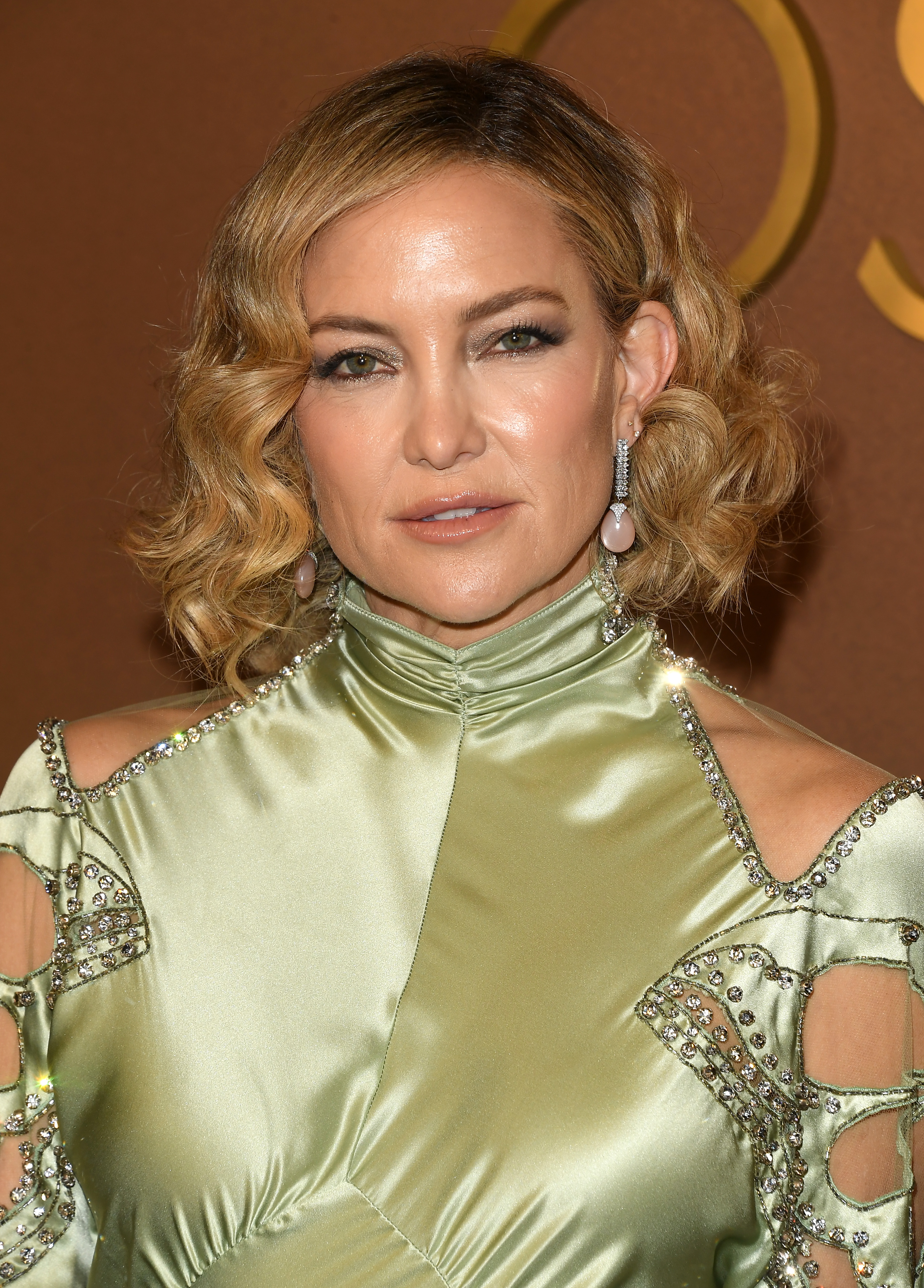 a close up red carpet photo of a white woman with short, blonde hair styled in curls. she&#039;s wearing a silky, light green gown with cutouts on the arms and shoulders