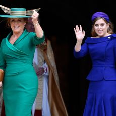 Sarah Ferguson wearing a green dress and matching hat smiling and standing next to Princess beatrice, who is dressed in a blue dress and headband and waving to the crowd on Princess Eugenie's wedding day