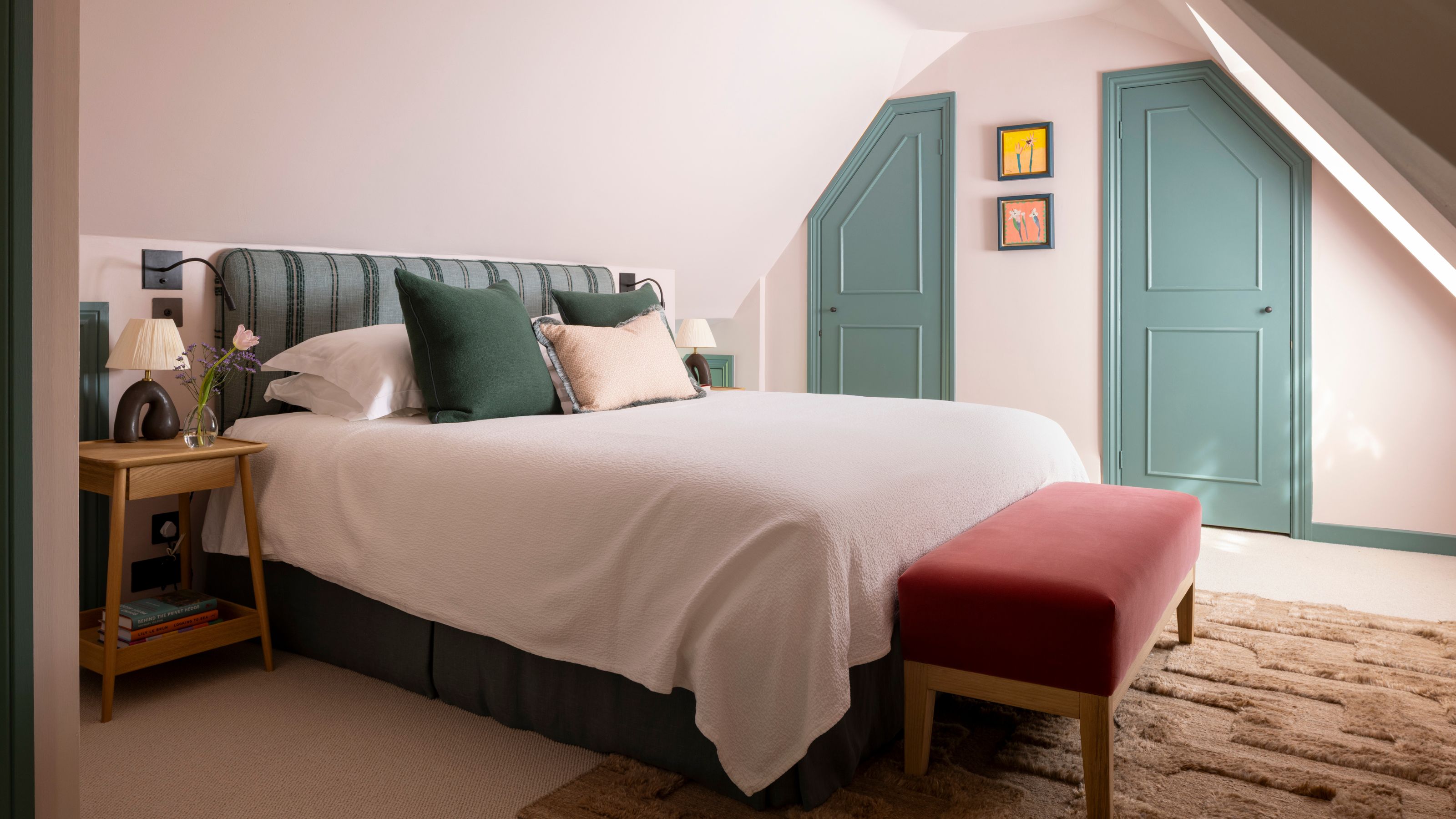 a loft bedroom with pale pink walls, green doors and trims, a striped headboard and dark green bed skirt with a red velvet bench and plush rug
