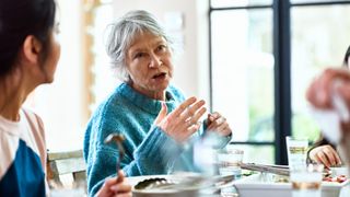 An older woman wearing a blue jumper is shown speaking at a dinner table. She is gesturing with her hands as she speaks. 