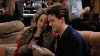 Maggie Wheeler and Matthew Perry in Friends sitting in Central Perk on the couch looking at something together.