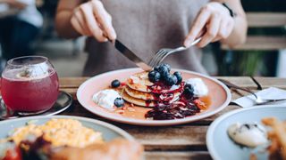 Close up of young woman sitting at dining table eating pancakes with blueberries and whipped cream in cafe, with English breakfast and french fries served on the dining table. Eating out lifestyle - stock photo