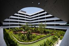 An open space courtyard with a manicured garden (with flowers and trees) in the center of a multi story grey steel building. Photographed curing the day from a parapet on the 2nd floor 