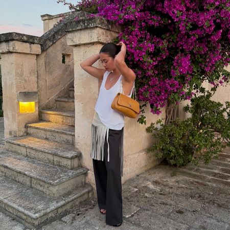 Creator @sasha.mei wearing black trousers and a fringed white top in a summery destination 