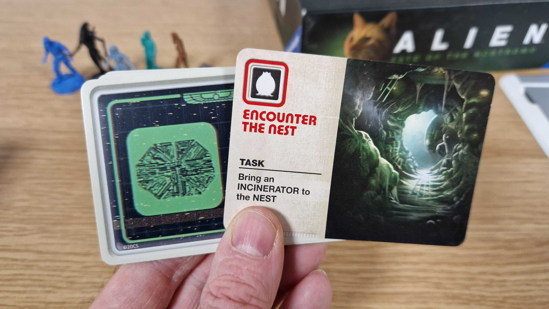 A hand holds up cards from Alien: Fate of the Nostromo in front of a wooden table with miniatures and the box on it