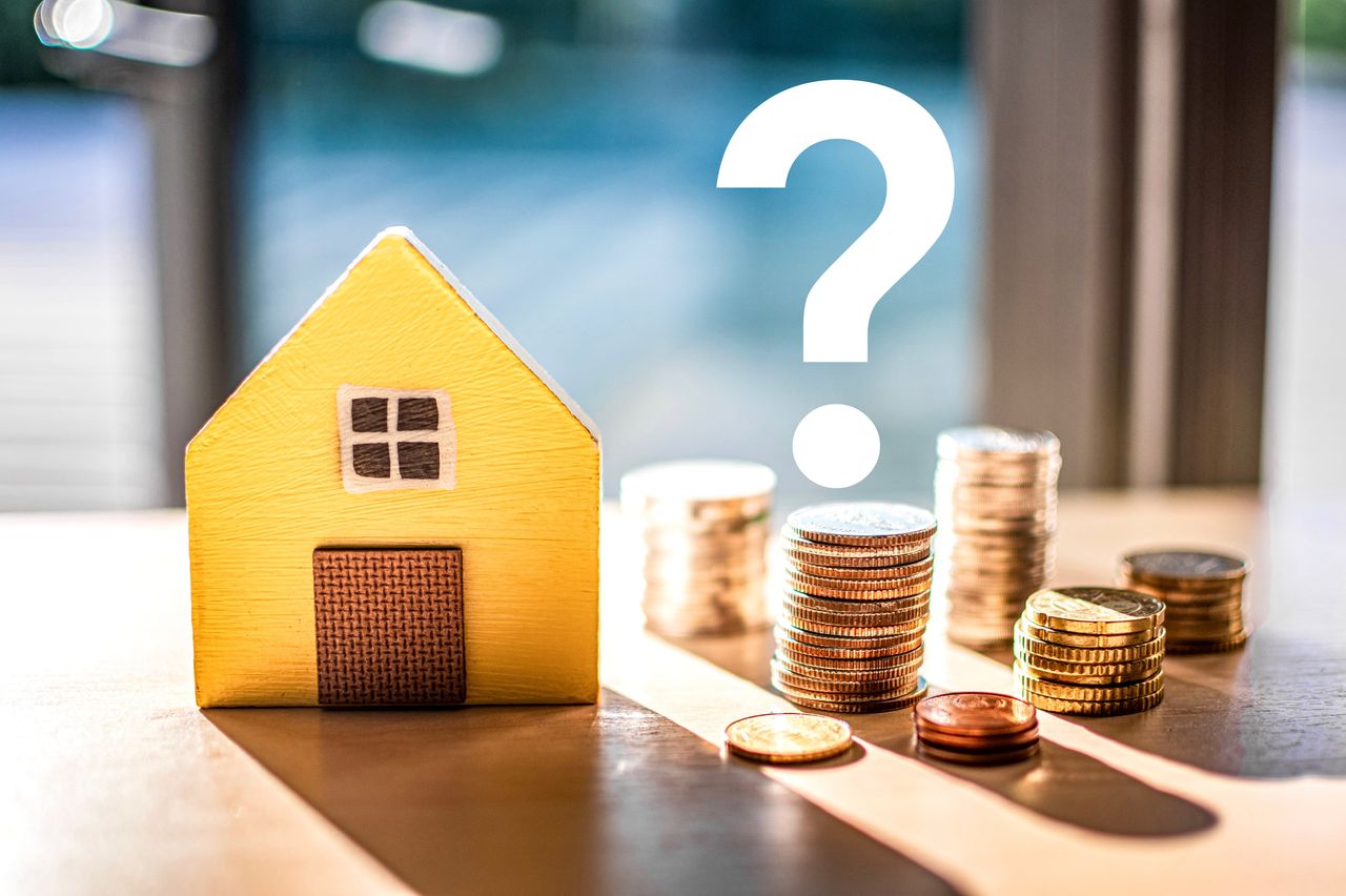 Hidden expenses of homebuying concept. Model wooden house sits next to a stack of coins, over which is a question mark. 