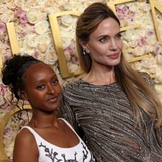 Angelina Jolie in a glittery black gown and her daughter Zahara standing in front of a rose covered Golden Globes step and repeat