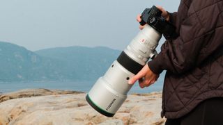 XF 500mm f/5.6 R LM OIS WR lens attached to a camera and held in front of a person outside on a rocky cliffside with mountains in the background