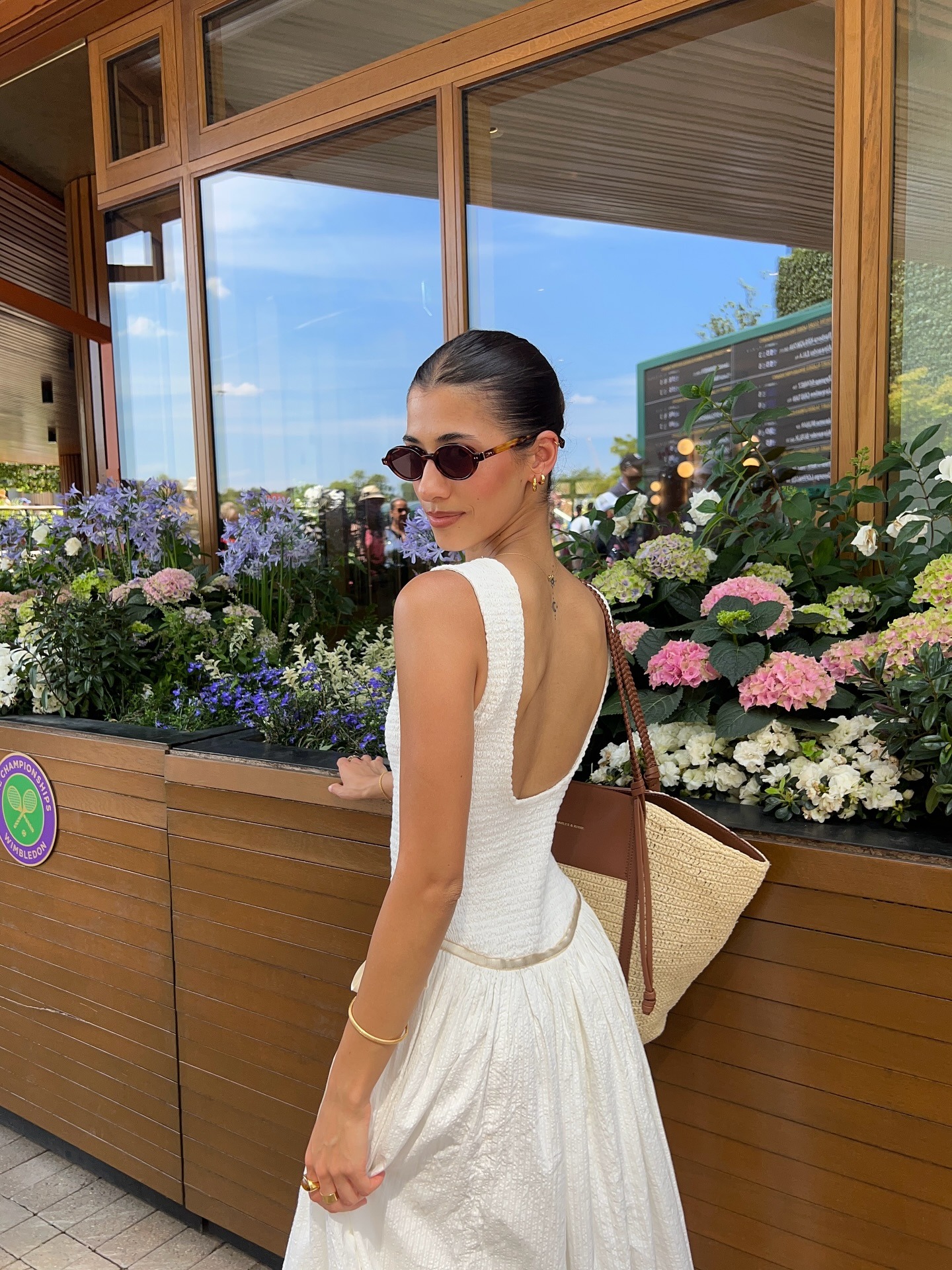 Fashion person Maya Fidel wears a Free People maxi dress, Miu Miu sungalsses and a raffia tote bag. She is looking towards the camera, posed at Wimbledon in a recent image from her Instagram.