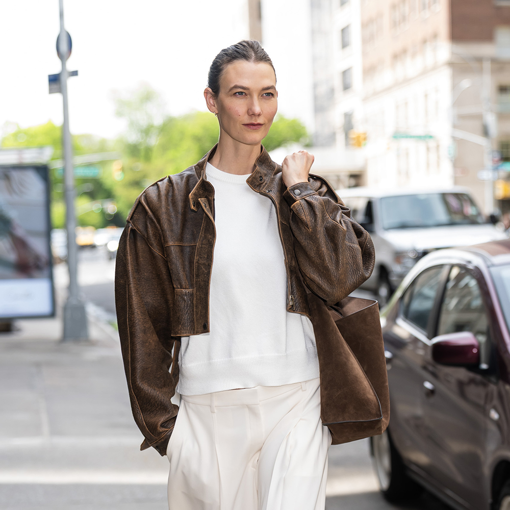 Karlie Kloss wearing a brown leather jacket, white tee, white pants, and brown suede flats