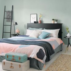 Small bedroom with double bed with black bedhead, pink, green and grey bedding, grey painted floorboards with a rug and retro suitcases at the foot of the bed.