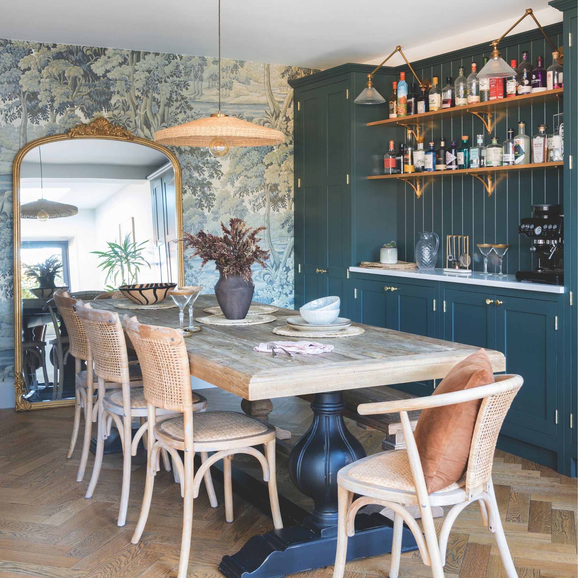 Large mixed material dining table with rattan chairs in a dining room with pattered wallpaper, large ornate mirror and built-in teal cabinetry for storage and a home bar