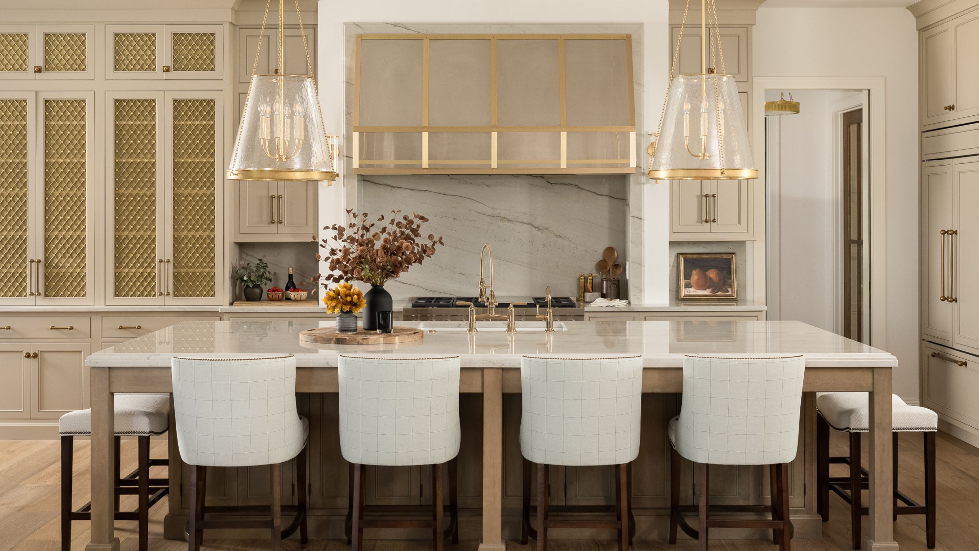 A luxurious kitchen view showing a large marble-topped island with four white barstools, centered under two large glass pendant lights and a custom gold-trimmed range hood.
