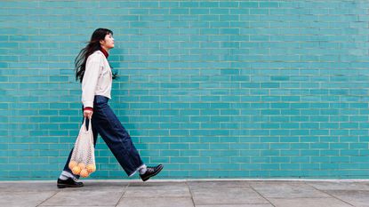 A young woman in trendy jeans walks carrying a mesh bag of oranges.