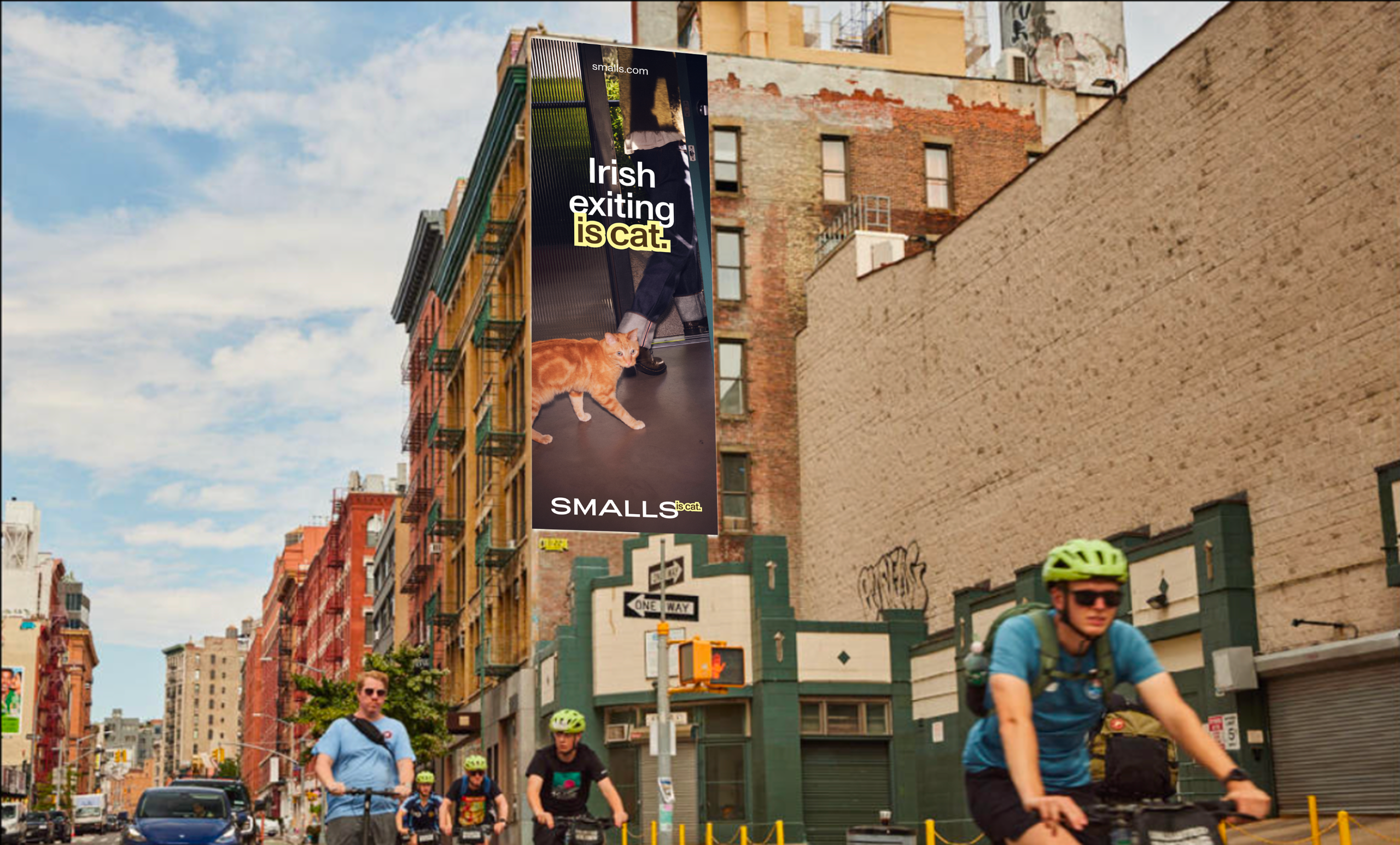 Mock-up of a tall Smalls &amp;quot;Irish exiting is cat&amp;quot; poster mounted on a New York City street corner, showing a ginger cat slipping past someone's feet, with cyclists passing below.