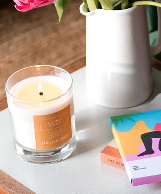 A white candle with a brown label on a white and wooden surface with colorful books and a white vase next to it