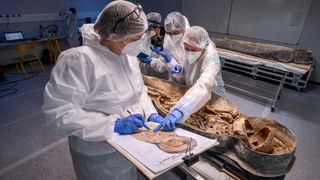 Four scientists in scrubs are in a room with a coffin on a table. The coffin is open and has a skeleton inside.