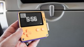 The Playdate handheld console in hand on an airplane with a tray table in its upright position in the background