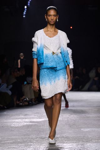 An S/S 25 model wearing a blue and white skirt set with a brooch in the center at the Bottega Veneta show.