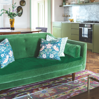 Open plan living area with pale pink walls, green velvet sofa on floral pattern rug, retro dining table and green kitchen units. Mid-century style mixed with ice cream shades for soothing seaside living.