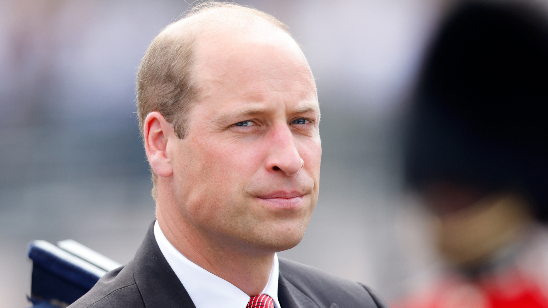 Prince William wears a black suit with a red tie and looks serious