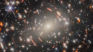 a field of galaxies in space, dominated by an enormous, bright-white elliptical galaxy that is the core of a massive galaxy cluster, surrounded by short, curved, glowing red lines, which are images of distant background galaxies magnified and warped by gravitational lensing.