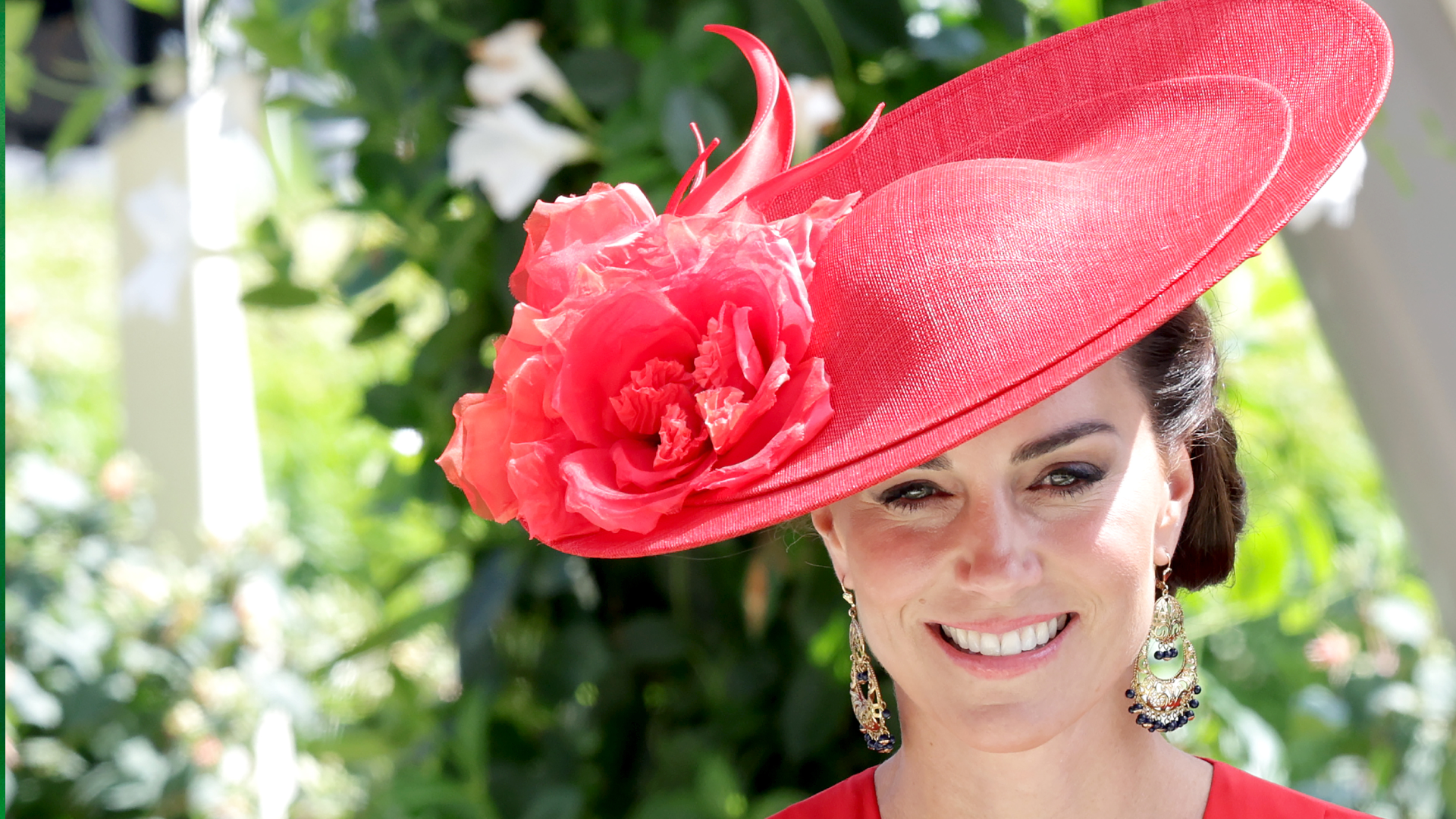 Kate Middleton wearing a red hat with flowers and dangling earrings smiling in front of flowers