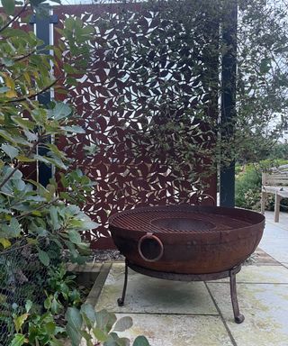 A dark brown metal privacy screen with a matching circular fire pit next to it and a green leafy bush in front of it